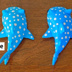 May include: Two whale shark toys, one light blue and one sea blue, both with white spots. The toys are lying on a wooden surface. The text 'Light Blue' is below the light blue whale shark and the text 'Sea Blue' is below the sea blue whale shark.