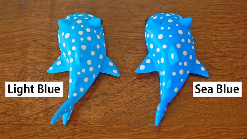 May include: Two whale shark toys, one light blue and one sea blue, both with white spots. The toys are lying on a wooden surface. The text 'Light Blue' is below the light blue whale shark and the text 'Sea Blue' is below the sea blue whale shark.