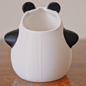 Op de afbeelding: Witte keramische panda-vormige plantenpot met zwarte oren en ogen. De pot heeft een rond lichaam en een brede opening aan de bovenkant.