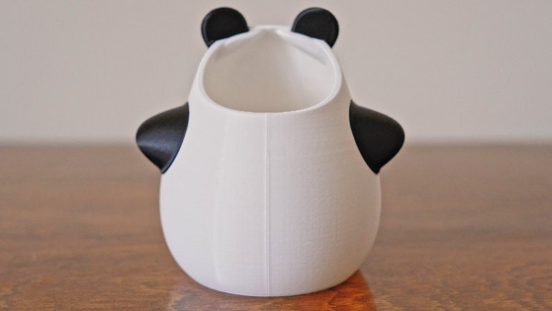 Op de afbeelding: Witte keramische panda-vormige plantenpot met zwarte oren en ogen. De pot heeft een rond lichaam en een brede opening aan de bovenkant.