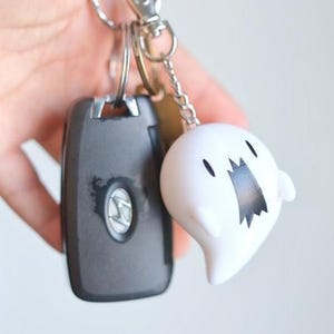 May include: A set of keys with a car key fob and a white ghost-shaped keychain charm. The ghost charm has a black mouth and eyes. The car key fob is dark gray with a silver emblem.