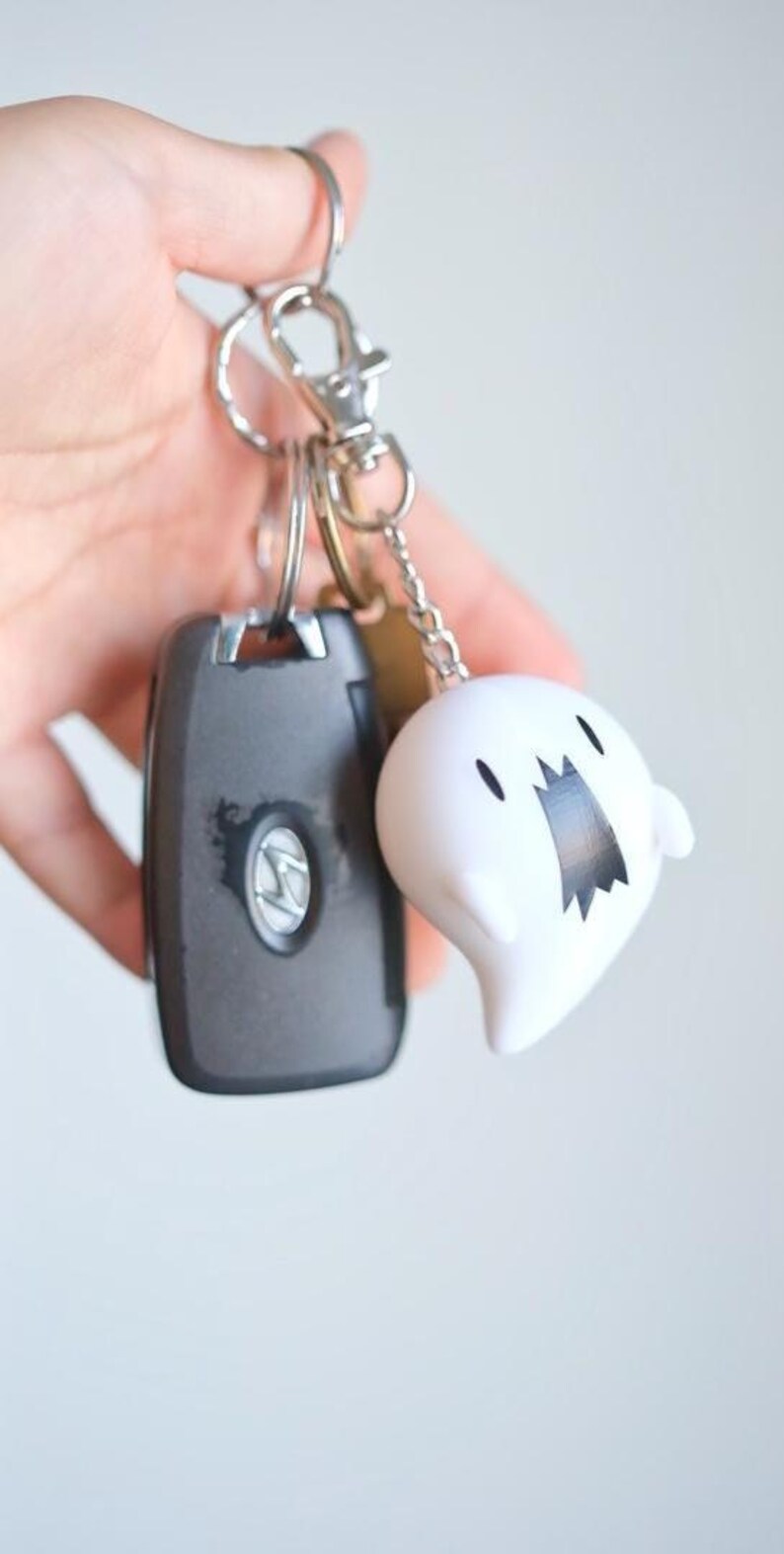 May include: A set of keys with a car key fob and a white ghost-shaped keychain charm. The ghost charm has a black mouth and eyes. The car key fob is dark gray with a silver emblem.