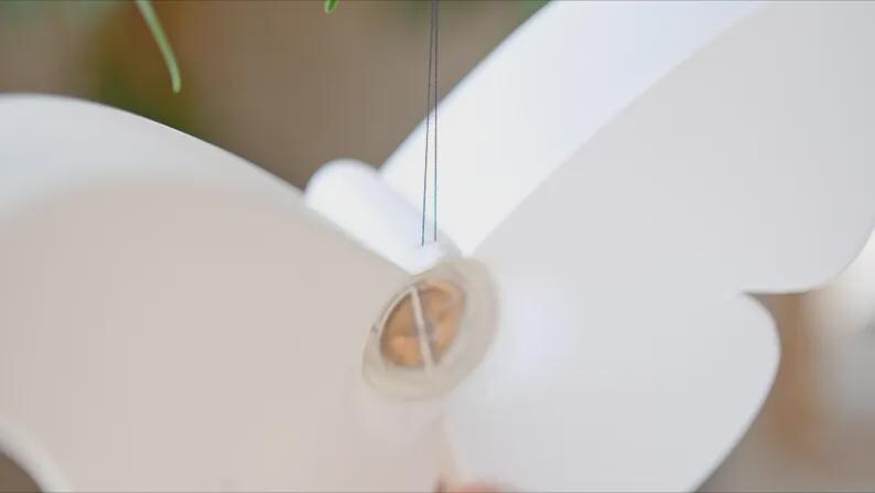 May include: A white paper butterfly with a circular, transparent center. The butterfly is suspended by thin, dark strings, suggesting it may be a mobile or decorative element. The wings are large and smooth.