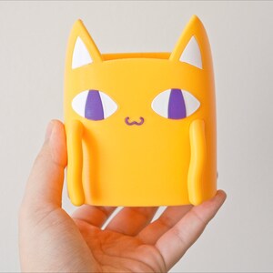 Peut inclure: Un porte-crayons en forme de chat jaune avec des yeux violets et des oreilles blanches.