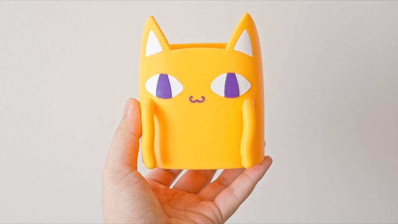 Peut inclure: Un porte-crayons en forme de chat jaune avec des yeux violets et des oreilles blanches.