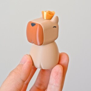 May include: A small, beige, cartoon dog figurine wearing a gold crown. The dog has a brown nose and black eyes.