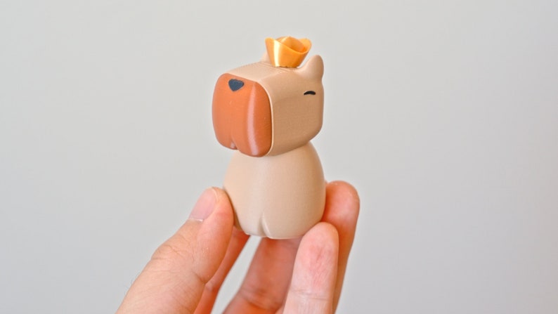 May include: A small, beige, cartoon dog figurine wearing a gold crown. The dog has a brown nose and black eyes.