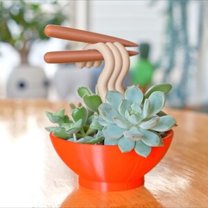 May include: A whimsical succulent planter shaped like a ramen bowl, with a vibrant orange exterior. The bowl holds green succulents and is topped with beige noodles and brown chopsticks. The planter sits on a wooden table.