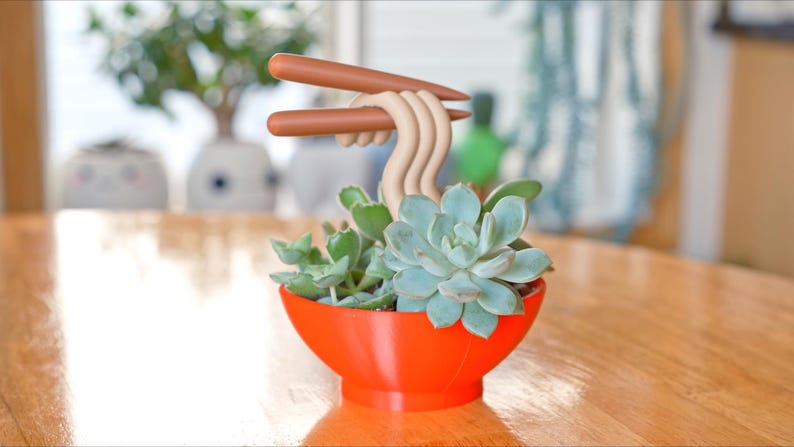 May include: A whimsical succulent planter shaped like a ramen bowl, with a vibrant orange exterior. The bowl holds green succulents and is topped with beige noodles and brown chopsticks. The planter sits on a wooden table.