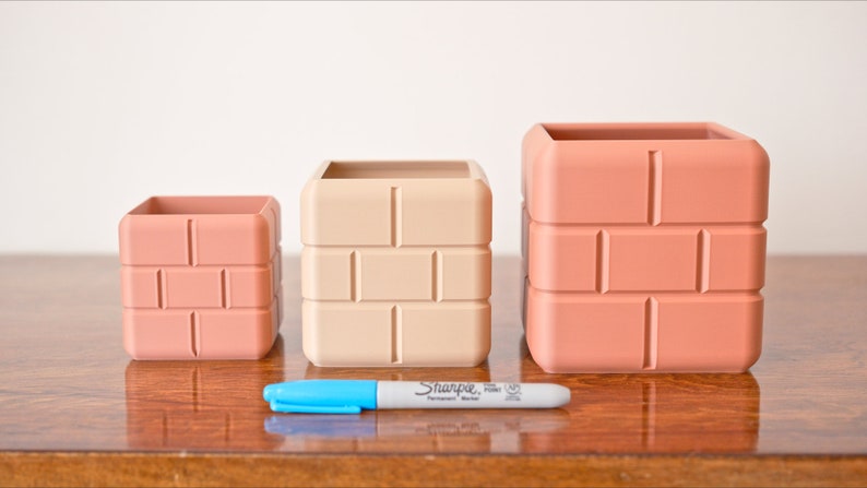 Pu&ograve; includere: Tre vasi quadrati di diverse dimensioni in terracotta con un motivo a mattoni. I vasi sono disposti dal pi&ugrave; piccolo al pi&ugrave; grande. Un pennarello blu &egrave; davanti ai vasi.