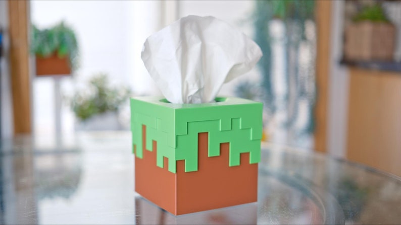 A cube tissue box cover inspired by the dirt grass block from the popular video game, Minecraft, is on a kitchen table with plants in the background. It has bright green pixelated grass at the top half and the bottom half is brown.