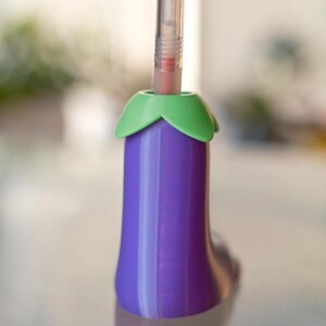 May include: A purple eggplant-shaped pen holder with a green top. A clear pen with a red clip is inserted into the holder.