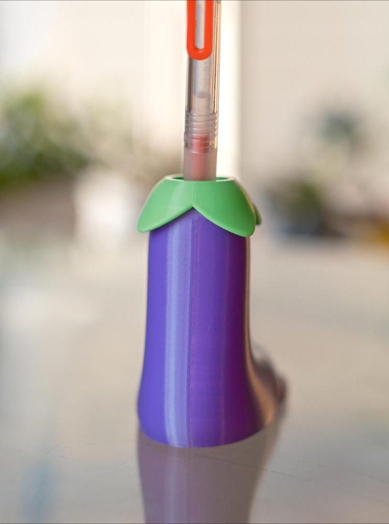 May include: A purple eggplant-shaped pen holder with a green top. A clear pen with a red clip is inserted into the holder.