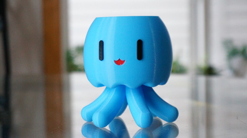 May include: A light blue 3D printed planter shaped like a smiling octopus with black eyes and a red mouth. The planter has six tentacles.