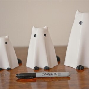 May include: Three white ceramic ghost figurines with black eyes. The ghosts are different sizes and are arranged in a row on a wooden surface.