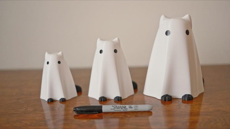 May include: Three white ceramic ghost figurines with black eyes. The ghosts are different sizes and are arranged in a row on a wooden surface.