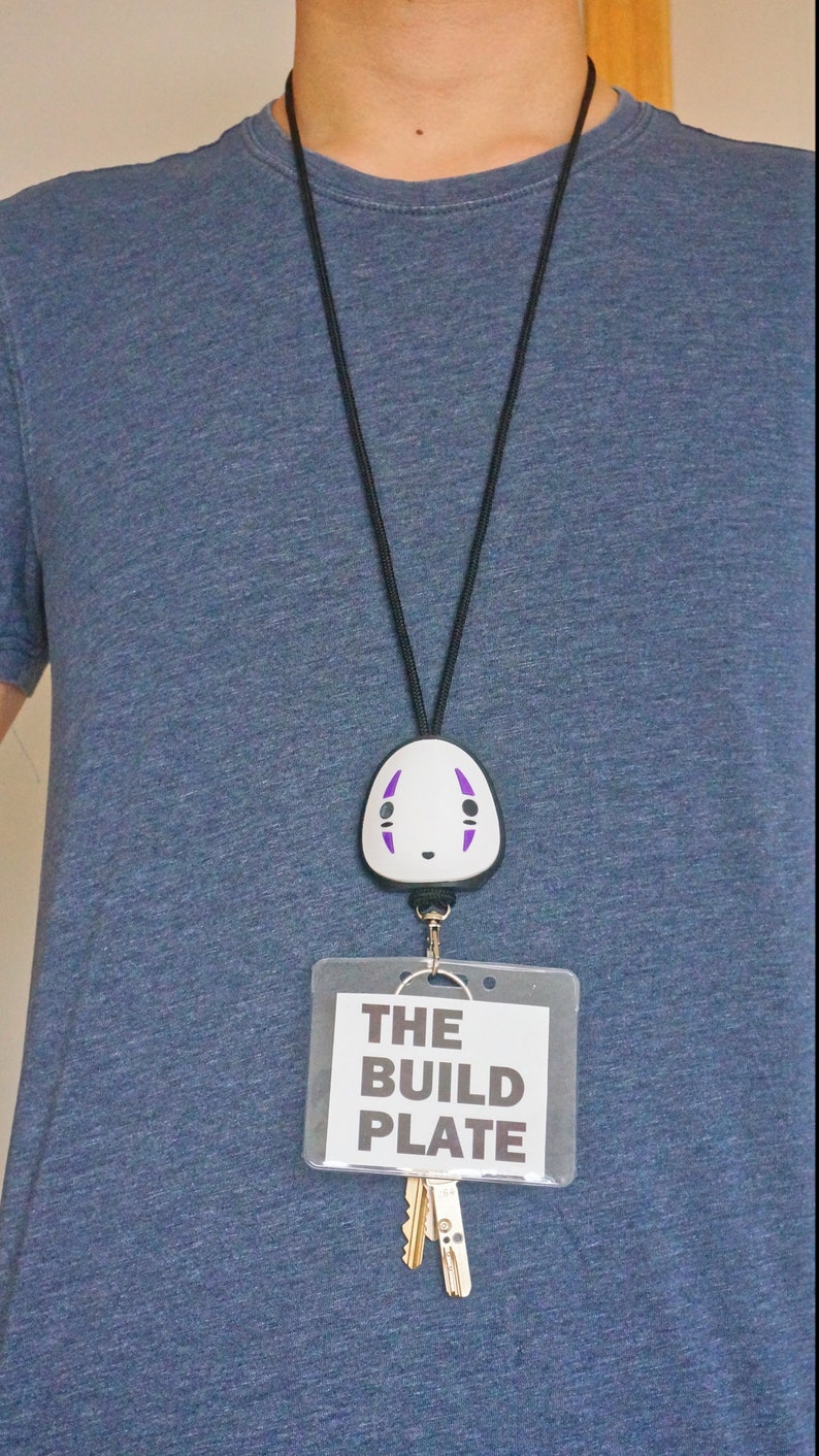 May include: A black lanyard with a white and purple No-Face character badge reel. Below the badge reel is a clear plastic card holder with the text "THE BUILD PLATE" and two keys.