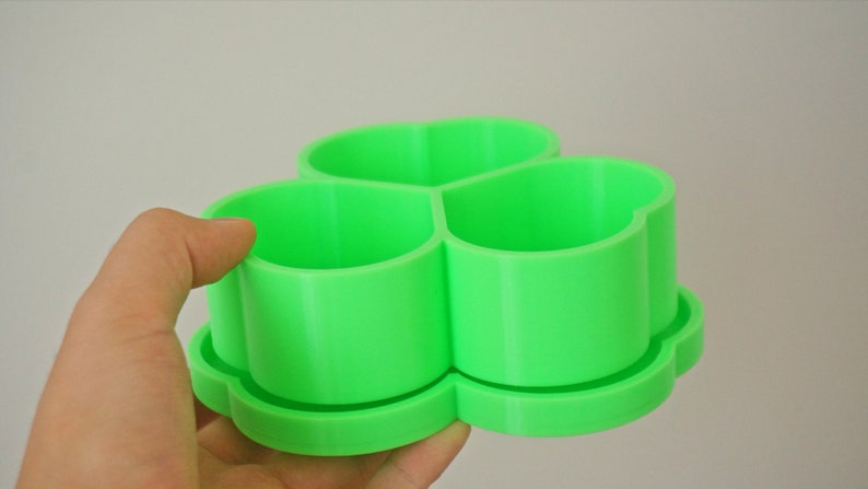 May include: A bright green, three-sectioned container with a clover-shaped base. The container is made of a smooth, solid material and is designed to hold small items. The design is simple and modern, suitable for various uses.