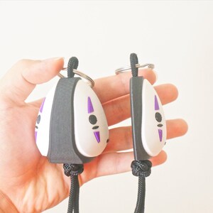 May include: Two No-Face keychains. Each keychain features a white face with purple markings and a black border. The keychains are attached to a black lanyard and a silver keyring. The keychains are held in a hand.