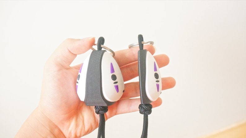 May include: Two No-Face keychains. Each keychain features a white face with purple markings and a black border. The keychains are attached to a black lanyard and a silver keyring. The keychains are held in a hand.