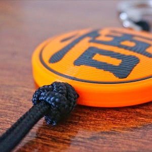Puede incluir: Primer plano de un llavero naranja con un car&aacute;cter chino negro. El llavero est&aacute; unido a un cord&oacute;n negro con un nudo.