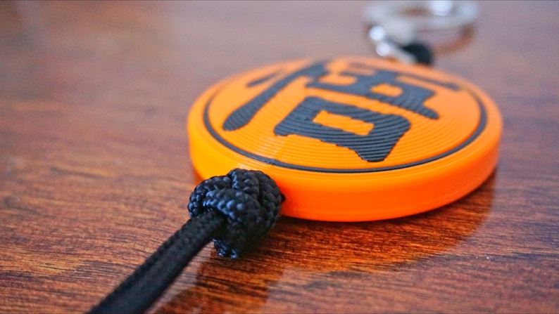 Puede incluir: Primer plano de un llavero naranja con un car&aacute;cter chino negro. El llavero est&aacute; unido a un cord&oacute;n negro con un nudo.