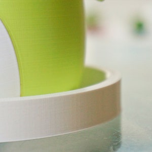Puede incluir: Una maceta blanca y verde con un platillo blanco. La maceta tiene una superficie lisa y texturizada.