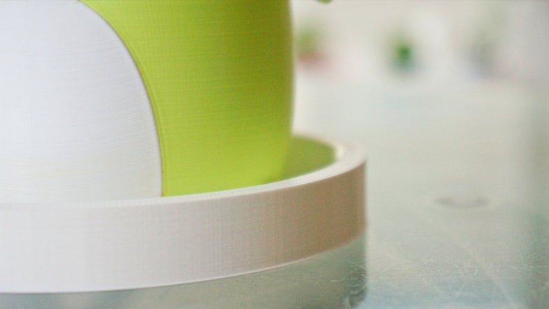 Puede incluir: Una maceta blanca y verde con un platillo blanco. La maceta tiene una superficie lisa y texturizada.