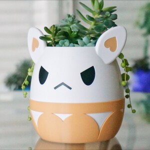 May include: White ceramic planter shaped like a cartoon animal with a grumpy expression. The planter has orange accents and is filled with green succulents.