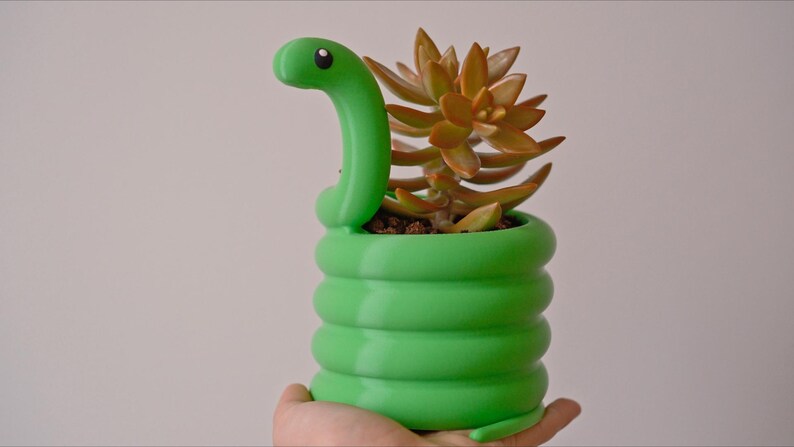 May include: A green snake-shaped planter holding a succulent plant. The planter is a vibrant green with a coiled body and a snake head. The succulent has orange-brown leaves.