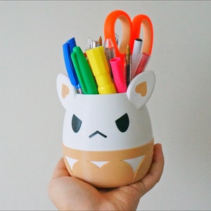 May include: White ceramic pencil holder shaped like a cartoon animal with brown ears and a brown bottom. The holder has a black angry face and is filled with colourful pens, pencils, and a pair of orange scissors.