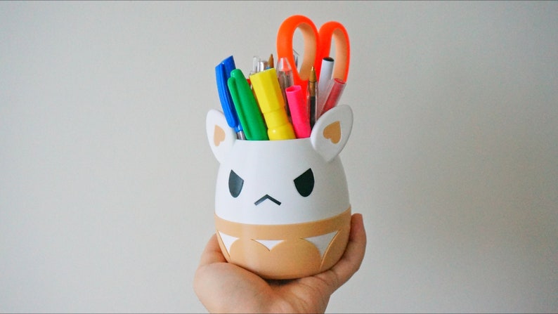 May include: White ceramic pencil holder shaped like a cartoon animal with brown ears and a brown bottom. The holder has a black angry face and is filled with colourful pens, pencils, and a pair of orange scissors.