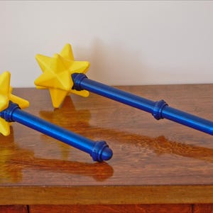 May include: Two blue and yellow star-shaped wands rest on a wooden surface. The wands have a yellow starburst at the top, a blue handle, and a blue base. The wands are likely props or decorative items.