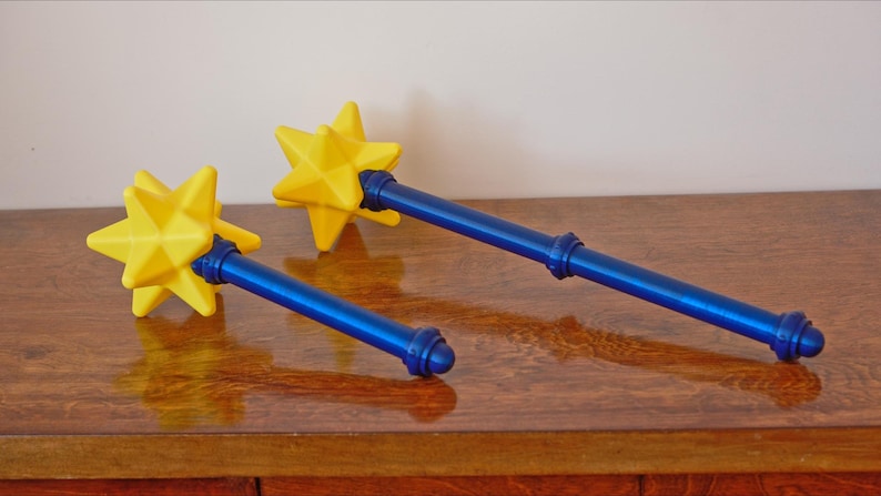 May include: Two blue and yellow star-shaped wands rest on a wooden surface. The wands have a yellow starburst at the top, a blue handle, and a blue base. The wands are likely props or decorative items.
