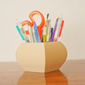 May include: A beige, two-toned pen holder filled with various pens, pencils, and scissors. The holder has a rounded shape with a flat base and is sitting on a wooden surface. The scissors are orange.