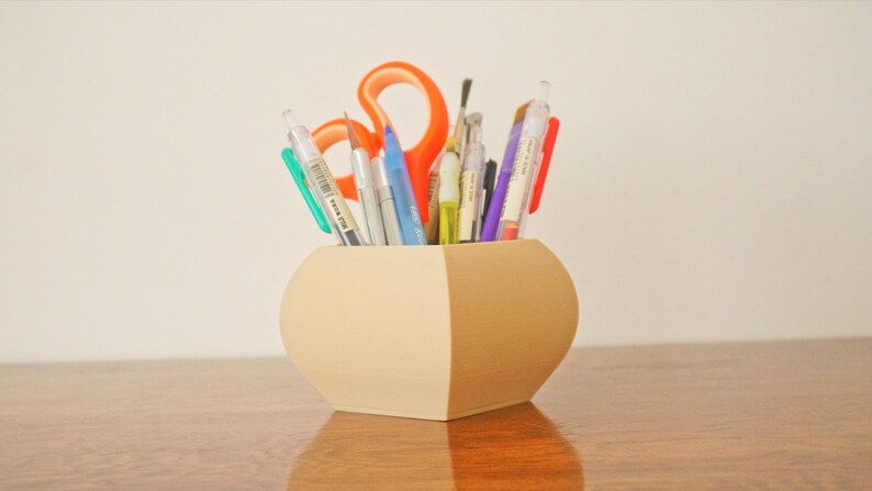 May include: A beige, two-toned pen holder filled with various pens, pencils, and scissors. The holder has a rounded shape with a flat base and is sitting on a wooden surface. The scissors are orange.