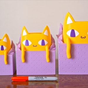 Peut inclure: Trois pots de fleurs en forme de chat jaunes avec des yeux violets et une bo&icirc;te violette &agrave; motif de points. Les pots sont de tailles diff&eacute;rentes et sont dispos&eacute;s du plus petit au plus grand.