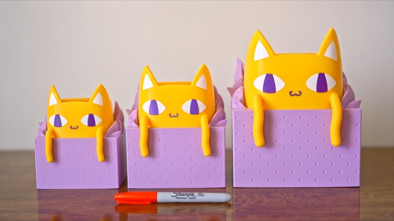 Peut inclure: Trois pots de fleurs en forme de chat jaunes avec des yeux violets et une bo&icirc;te violette &agrave; motif de points. Les pots sont de tailles diff&eacute;rentes et sont dispos&eacute;s du plus petit au plus grand.