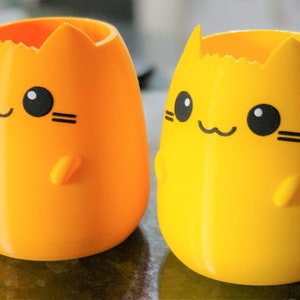 May include: Two cat-shaped pen holders in bright orange and yellow. Each has a smiling face with black eyes, whiskers, and a small paw. The holders are egg-shaped with a jagged top, and are made of a smooth plastic.