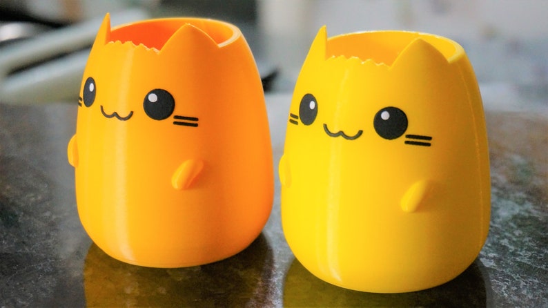 May include: Two cat-shaped pen holders in bright orange and yellow. Each has a smiling face with black eyes, whiskers, and a small paw. The holders are egg-shaped with a jagged top, and are made of a smooth plastic.