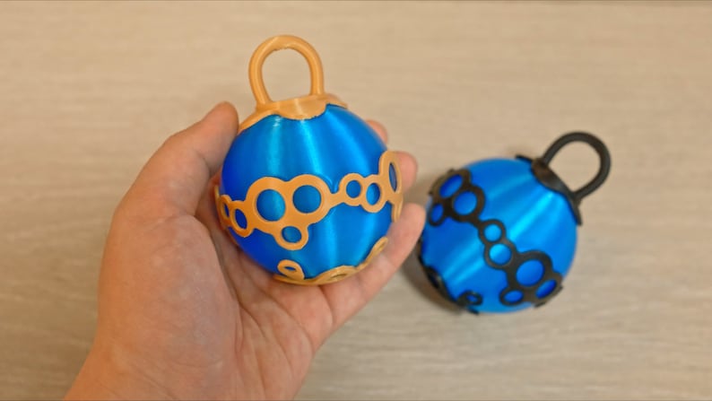 May include: Two blue spherical ornaments with decorative patterns and loop handles. One ornament has gold accents, the other black. The ornaments are likely for decoration or crafts.