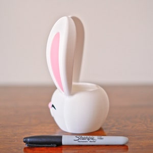 May include: White ceramic bunny planter with pink ears. The planter is shaped like a bunny with a round body and long ears. The bunny has a small, closed mouth and a small, black nose.