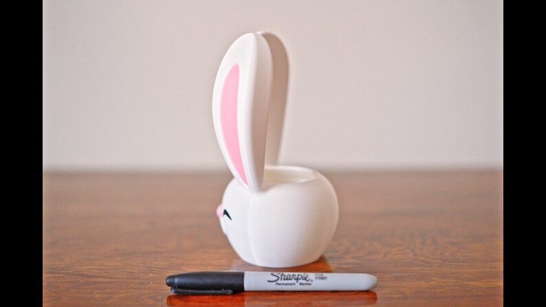 May include: White ceramic bunny planter with pink ears. The planter is shaped like a bunny with a round body and long ears. The bunny has a small, closed mouth and a small, black nose.