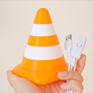 May include: Orange and white striped traffic cone-shaped lamp with a USB cable and on/off switch. The lamp is held in a hand against a neutral background. The lamp is a fun, decorative item.