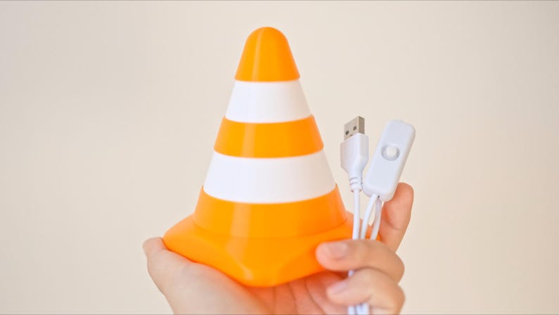 May include: Orange and white striped traffic cone-shaped lamp with a USB cable and on/off switch. The lamp is held in a hand against a neutral background. The lamp is a fun, decorative item.