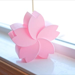 May include: A pink paper flower with five petals, resembling a cherry blossom, sits on a white windowsill.