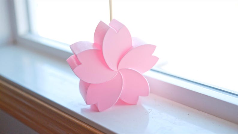 May include: A pink paper flower with five petals, resembling a cherry blossom, sits on a white windowsill.