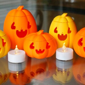 May include: Five orange and yellow jack-o'-lantern candle holders with carved faces. Each pumpkin has a small LED tea light inside. The pumpkins are arranged on a glass table.