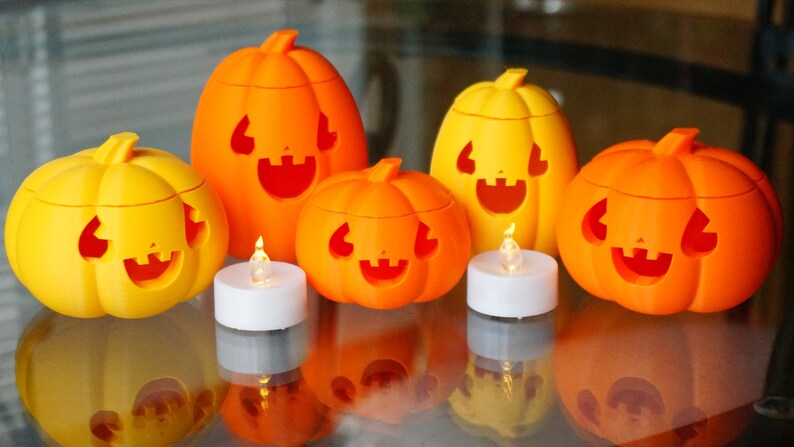 May include: Five orange and yellow jack-o'-lantern candle holders with carved faces. Each pumpkin has a small LED tea light inside. The pumpkins are arranged on a glass table.
