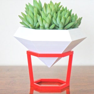 May include: A white geometric diamond-shaped planter with a succulent plant inside, sitting on a red metal stand.